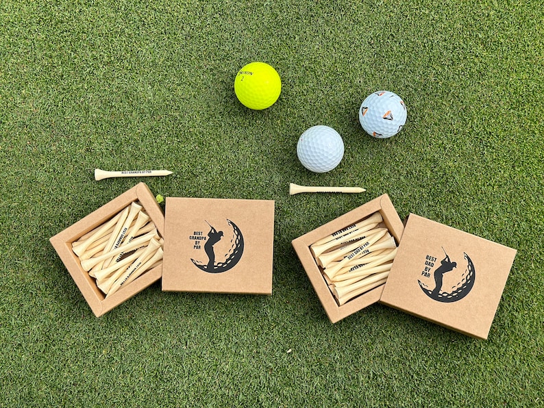 Puede incluir: Juego de regalo con tem&aacute;tica de golf que incluye pelotas de golf, tees de madera y cajas con la silueta de un golfista. Las cajas y los tees tienen el texto "BEST GRANDPA BY PAR". Las pelotas son blancas, amarillas y estampadas, sobre una superficie verde.