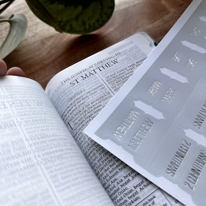 May include: A close-up of a white sticker sheet with silver lettering. The stickers are for different books of the Bible, including Matthew, Mark, Luke, and 2 Corinthians. The stickers are designed to be placed on the spines of books.