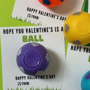 May include: Three Valentine's Day cards with a soccer ball theme. Each card has a different colored soccer ball attached to it. The text on the cards reads "Happy Valentine's Day" and "Hope you Valentine's is a ball".