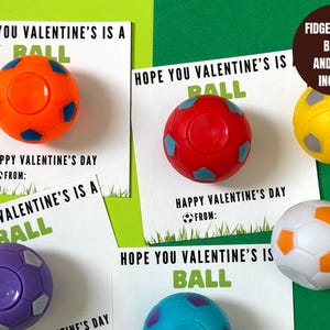 May include: Four Valentine's Day cards with the text "Hope you Valentine's is a ball" and a small soccer ball attached to each card. The soccer balls are different colors: orange, red, purple, and blue. The text "Fidget Soccer Balls and Cards Included" is on a brown label.