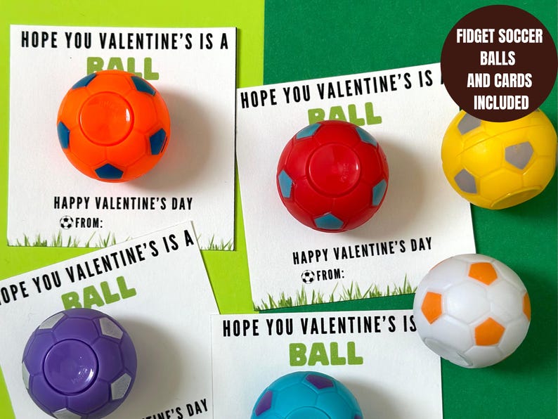 May include: Four Valentine's Day cards with the text "Hope you Valentine's is a ball" and a small soccer ball attached to each card. The soccer balls are different colors: orange, red, purple, and blue. The text "Fidget Soccer Balls and Cards Included" is on a brown label.