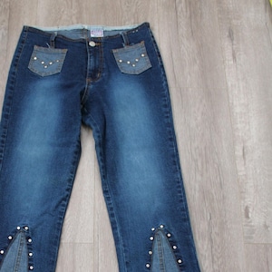 以下が含まれることがあります： 薄い色のウォッシュ加工が施された青いデニムのジーンズ。ポケットと縫い目にラインストーンの装飾が施されています。ジーンズはブーツカットスタイルで、裾はフリンジになっています。