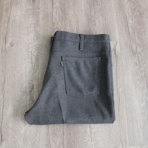 Puede incluir: Un pantalón gris doblado sobre una superficie clara con vetas de madera. El pantalón tiene un solo bolsillo trasero y una pequeña etiqueta. La tela parece ser un material suave y tejido. El pantalón es de color liso y neutro.
