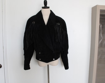 May include: Black suede jacket with fringe detailing on the sleeves and shoulders. The jacket has a notched collar, a wrap-style front, and two silver-tone buttons. The jacket is displayed on a mannequin.