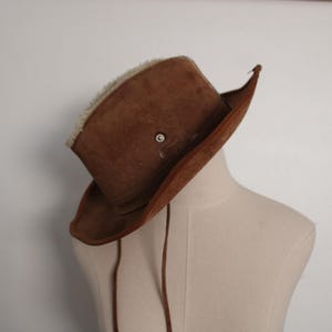 May include: A brown suede cowboy hat with a faux fur band. The hat has a wide brim and a pointed crown.