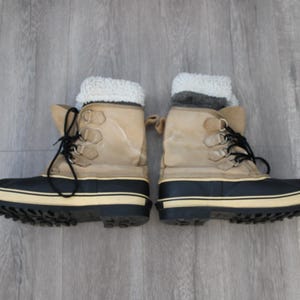 May include: A pair of tan winter boots with black trim and white soles. The boots have a faux fur lining and are tied with black laces.