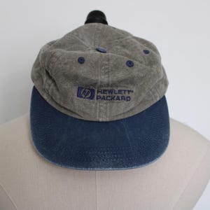 Puede incluir: Una gorra de béisbol gris y azul con el logotipo de Hewlett Packard bordado en la parte delantera.