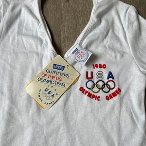 May include: White t-shirt with a red, white, and blue "1980 USA Olympic Games" graphic on the front. The shirt has a v-neck and short sleeves. A Levi's tag is attached to the shirt.