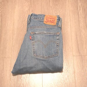 May include: Folded blue denim jeans with a classic five-pocket design. The jeans feature a brown leather patch with red lettering and a small red tag on the back pocket. The jeans are folded neatly on a light brown wooden surface.