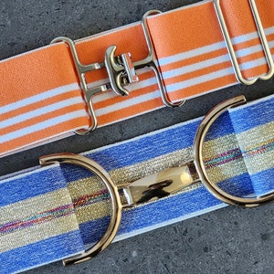 Pode incluir: Dois cintos el&aacute;sticos com estilos de fivela diferentes. Um cinto &eacute; laranja com listras brancas e uma fivela prateada. O outro cinto &eacute; azul com uma faixa dourada e uma fivela dourada.