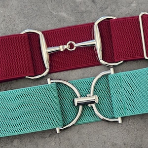 May include: Two elastic belts with silver-colored metal hardware. One belt is burgundy, and the other is teal. The belts feature a horse bit design and a rectangular buckle. The background is a neutral gray.
