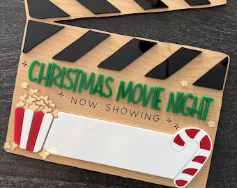 May include: A wooden movie clapperboard with black and green text that reads "CHRISTMAS MOVIE NIGHT NOW SHOWING." The clapperboard features a popcorn bucket and candy cane design.