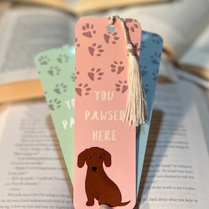 Peut inclure: Deux marque-pages rose et vert avec des empreintes de pattes et une illustration de teckel. Le marque-page rose dit "You Pawsed Here".
