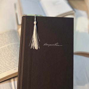 May include: A dark brown hardcover book with a white tassel bookmark. The book has the author's name, "Harper Lee", written in white on the front cover.
