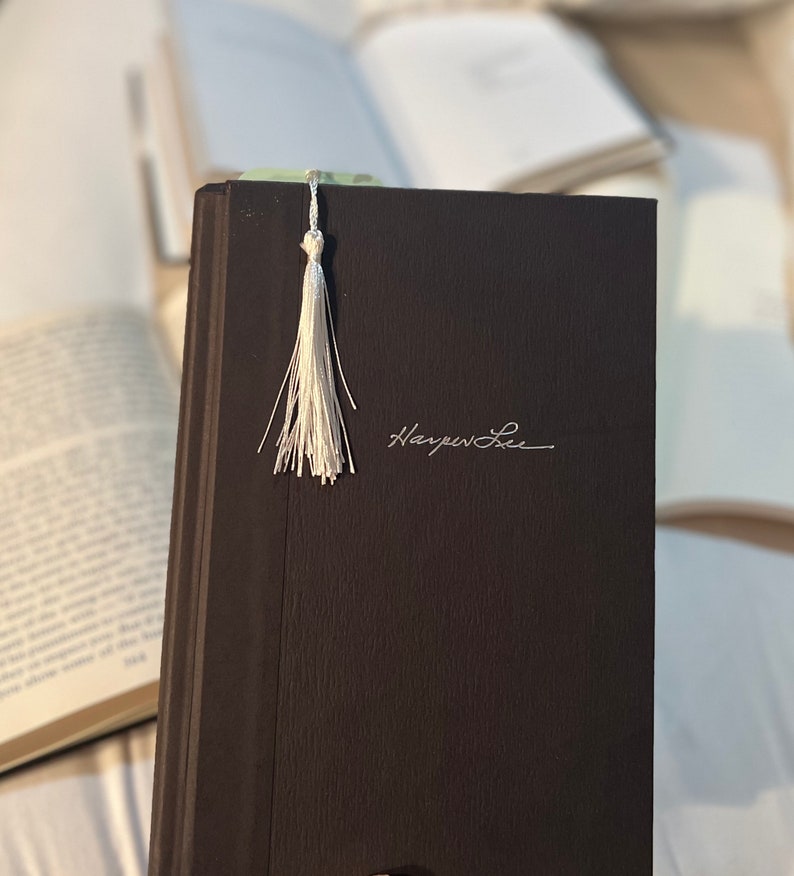May include: A dark brown hardcover book with a white tassel bookmark. The book has the author's name, "Harper Lee", written in white on the front cover.