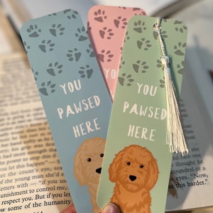 May include: Three colourful bookmarks with dog paw prints and the text "YOU PAWSED HERE." Each bookmark features a different background colour: light blue, pink, and green. A fluffy dog illustration is on each bookmark. One bookmark has a silver tassel.
