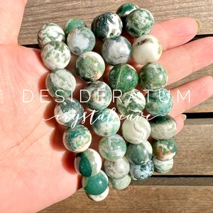 May include: A green and white agate gemstone bracelet with a variety of patterns. The bracelet is made of round beads and is strung on a clear elastic cord. The bracelet is on a hand, and the text "DESIDERATUM crystalcave" is visible in the background.