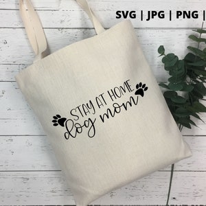 Peut inclure: Un sac fourre-tout en toile blanche avec du texte noir qui dit "Stay at home dog mom" avec deux empreintes de pattes de chaque c&ocirc;t&eacute; du texte.