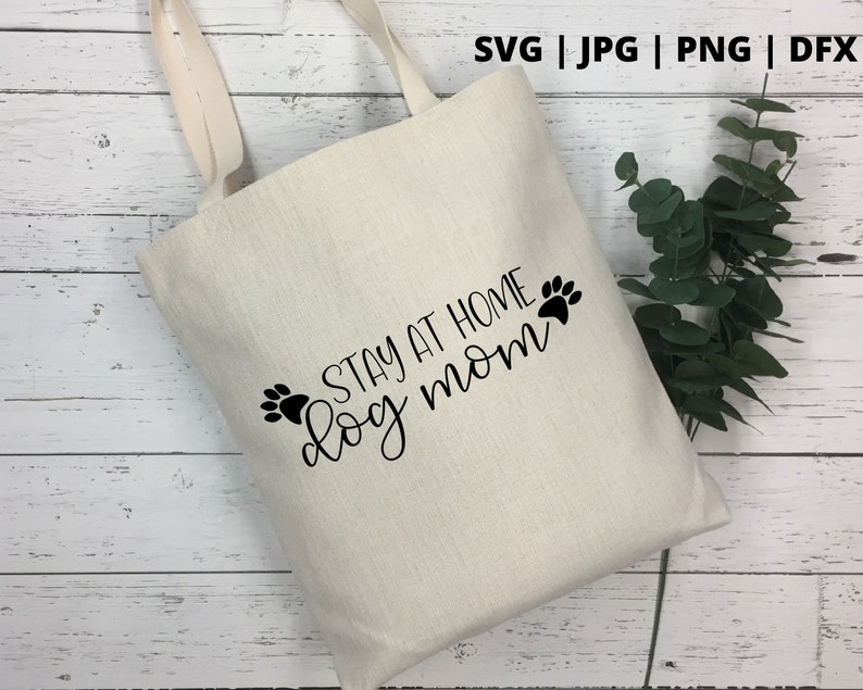 Peut inclure: Un sac fourre-tout en toile blanche avec du texte noir qui dit "Stay at home dog mom" avec deux empreintes de pattes de chaque c&ocirc;t&eacute; du texte.