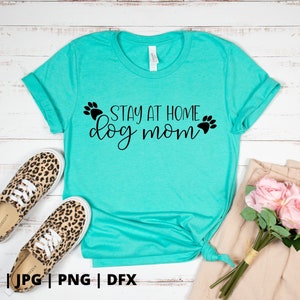 Peut inclure: T-shirt bleu turquoise avec le texte "Stay at Home Dog Mom" en lettres noires avec des empreintes de pattes de chaque c&ocirc;t&eacute; du texte.