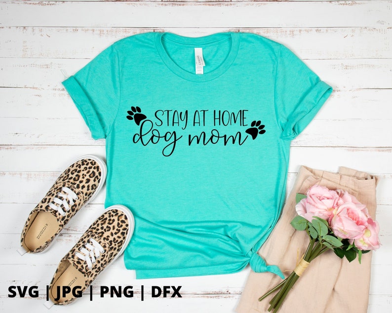 Peut inclure: T-shirt bleu turquoise avec le texte "Stay at Home Dog Mom" en lettres noires avec des empreintes de pattes de chaque c&ocirc;t&eacute; du texte.