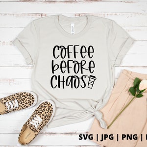 Op de afbeelding: Een wit T-shirt met zwarte tekst die "Coffee Before Chaos" zegt en een klein koffiekopje-icoon. Het shirt ligt op een wit houten oppervlak met een paar luipaardprint schoenen en een beige broek.