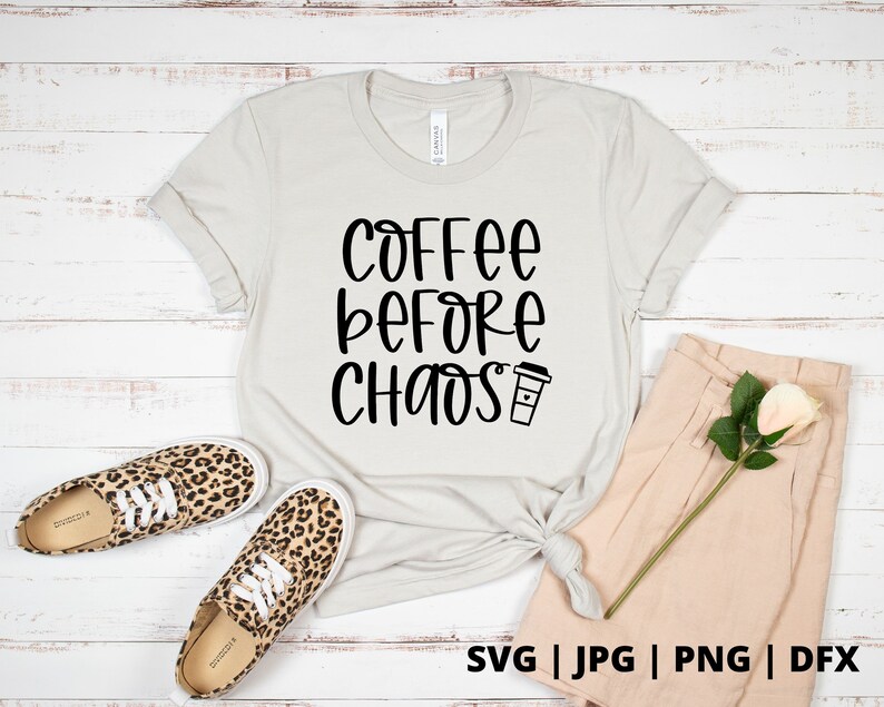 Op de afbeelding: Een wit T-shirt met zwarte tekst die "Coffee Before Chaos" zegt en een klein koffiekopje-icoon. Het shirt ligt op een wit houten oppervlak met een paar luipaardprint schoenen en een beige broek.