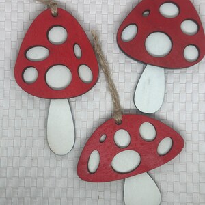 Op de afbeelding: Drie rode en witte houten paddenstoelversieringen met witte stelen. De paddenstoelen hebben een rustieke, handgeschilderde uitstraling.