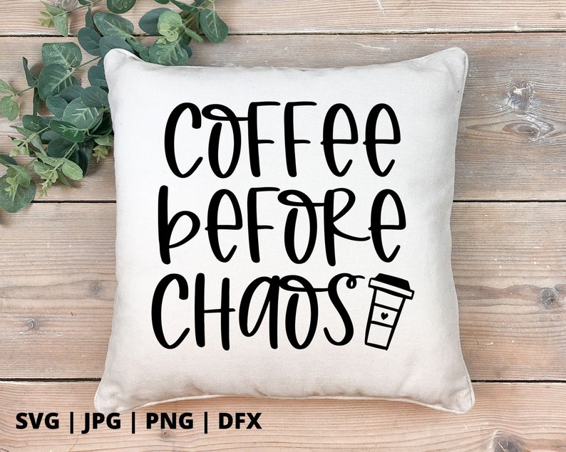 Op de afbeelding: Een wit kussen met zwarte tekst die "Coffee Before Chaos" luidt en een zwart koffiekopje-icoon.