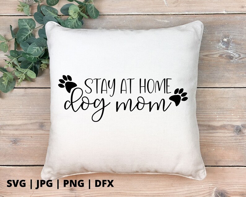 Peut inclure: Un oreiller blanc avec du texte noir qui dit "Stay at Home Dog Mom" avec des empreintes de pattes de chaque c&ocirc;t&eacute; du texte.
