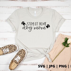 Peut inclure: Un t-shirt blanc avec le texte "Stay at Home Dog Mom" en lettres noires avec des empreintes de pattes de chaque c&ocirc;t&eacute; du texte. Le t-shirt est pos&eacute; sur un fond en bois blanc avec une paire de baskets l&eacute;opard et un short beige.