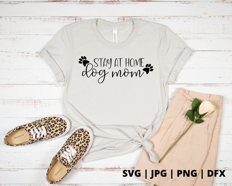 Peut inclure: Un t-shirt blanc avec le texte "Stay at Home Dog Mom" en lettres noires avec des empreintes de pattes de chaque c&ocirc;t&eacute; du texte. Le t-shirt est pos&eacute; sur un fond en bois blanc avec une paire de baskets l&eacute;opard et un short beige.