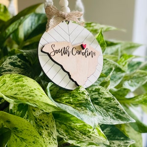 May include: A round wooden ornament featuring the outline of South Carolina, with the words "South Carolina" in a script font. A small red heart is placed on the map. The ornament hangs from a jute bow and wooden beads.