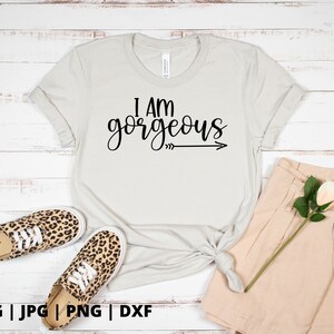 K&ouml;nnte beinhalten: Ein wei&szlig;es T-Shirt mit dem Text "I am gorgeous" in schwarzer Schrift mit einem Pfeil, der nach rechts zeigt. Das Shirt liegt auf einer wei&szlig;en Holzoberfl&auml;che mit einem Paar Leoparden-Sneakers und einer beigefarbenen Shorts.