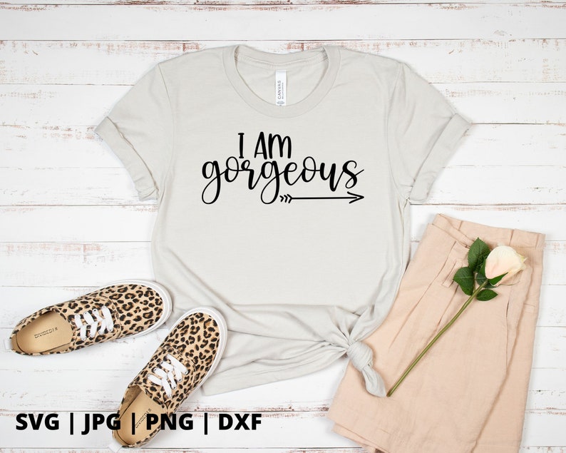 K&ouml;nnte beinhalten: Ein wei&szlig;es T-Shirt mit dem Text "I am gorgeous" in schwarzer Schrift mit einem Pfeil, der nach rechts zeigt. Das Shirt liegt auf einer wei&szlig;en Holzoberfl&auml;che mit einem Paar Leoparden-Sneakers und einer beigefarbenen Shorts.