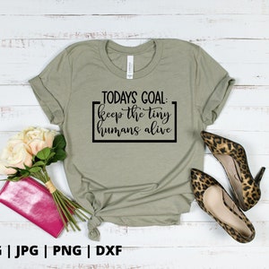 K&ouml;nnte beinhalten: Ein hellgr&uuml;nes T-Shirt mit dem Text "TODAYS GOAL: keep the tiny humans alive", der in Schwarz auf der Vorderseite aufgedruckt ist. Das Shirt liegt flach auf einer wei&szlig;en Holzoberfl&auml;che mit einem Strau&szlig; wei&szlig;er Rosen und einer rosa Clutch.