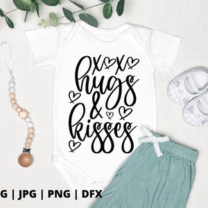 K&ouml;nnte beinhalten: Ein wei&szlig;er Baby-Strampler mit schwarzem Text, der "xox hugs & kisses" lautet, mit kleinen Herzen um den Text herum. Der Strampler liegt auf einer wei&szlig;en Oberfl&auml;che mit einem Paar wei&szlig;er Babyschuhe und einem Holz-Bei&szlig;ring.