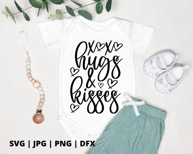 K&ouml;nnte beinhalten: Ein wei&szlig;er Baby-Strampler mit schwarzem Text, der "xox hugs & kisses" lautet, mit kleinen Herzen um den Text herum. Der Strampler liegt auf einer wei&szlig;en Oberfl&auml;che mit einem Paar wei&szlig;er Babyschuhe und einem Holz-Bei&szlig;ring.