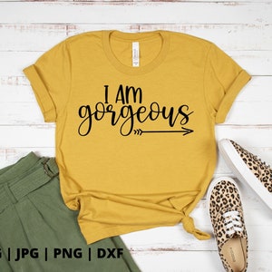 K&ouml;nnte beinhalten: Ein gelbes T-Shirt mit dem Text "I am gorgeous" in schwarzen Buchstaben mit einem Pfeil, der nach rechts zeigt. Das Shirt liegt auf einer wei&szlig;en Holzoberfl&auml;che mit einem Paar Leoparden-Schuhen.