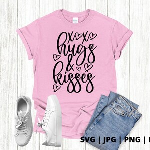 K&ouml;nnte beinhalten: Pinkes T-Shirt mit schwarzem Text, der "XOXO hugs & kisses" lautet, mit kleinen Herzen um den Text herum. Das Shirt liegt auf einem wei&szlig;en Holzgrund mit einem Paar wei&szlig;en Turnschuhen und einer blauen Jeans.