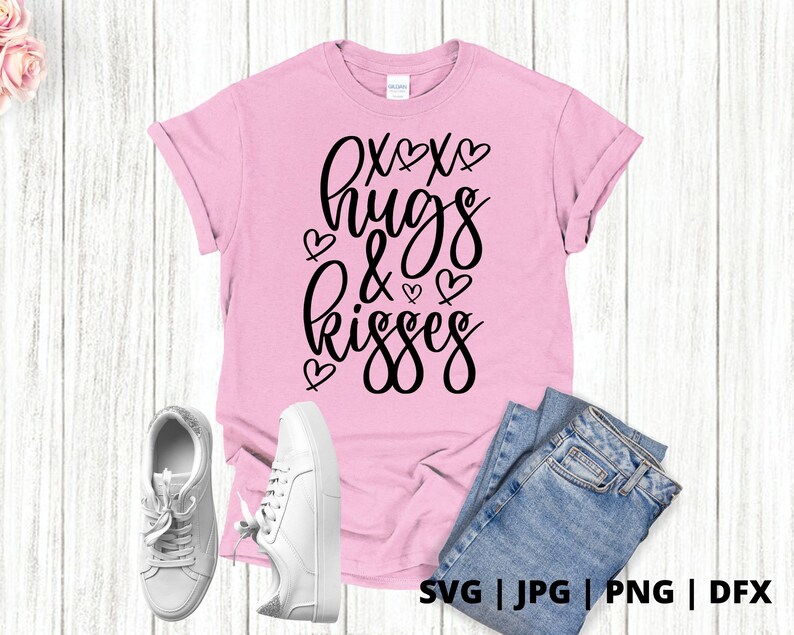 K&ouml;nnte beinhalten: Pinkes T-Shirt mit schwarzem Text, der "XOXO hugs & kisses" lautet, mit kleinen Herzen um den Text herum. Das Shirt liegt auf einem wei&szlig;en Holzgrund mit einem Paar wei&szlig;en Turnschuhen und einer blauen Jeans.