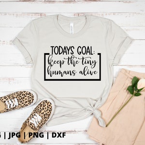 K&ouml;nnte beinhalten: Ein wei&szlig;es T-Shirt mit dem Text "TODAYS GOAL: keep the tiny humans alive", der in Schwarz auf der Vorderseite aufgedruckt ist. Das Shirt liegt auf einer Holzoberfl&auml;che mit einem Paar Leoparden-Schuhen und einer beigefarbenen Hose.