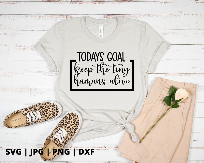 K&ouml;nnte beinhalten: Ein wei&szlig;es T-Shirt mit dem Text "TODAYS GOAL: keep the tiny humans alive", der in Schwarz auf der Vorderseite aufgedruckt ist. Das Shirt liegt auf einer Holzoberfl&auml;che mit einem Paar Leoparden-Schuhen und einer beigefarbenen Hose.