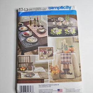 May include: Simplicity sewing pattern number 1343 for a table runner, placemats, and napkins. The pattern shows a variety of table settings with an autumn theme, including pumpkins, gourds, and plaid fabric. The pattern is designed and printed in the USA.