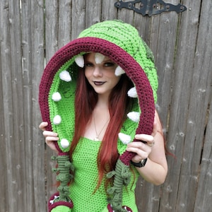 May include: A green crochet hooded costume with a large, open-mouthed design. The hood features a burgundy-colored mouth with white teeth and is accented with green and white details. The costume includes matching arm pieces and a green dress.