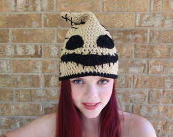 Puede incluir: Un gorro de punto de ganchillo con un diseño en negro y beige que presenta una cara cosida con una amplia sonrisa. El gorro lo lleva una persona con el pelo largo y rojo.