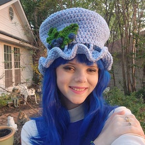 May include: A light blue crochet hat with a ruffled brim, adorned with a green leaf and blue berry embellishments. The hat is handmade and has a rounded crown. The hat is a whimsical accessory.