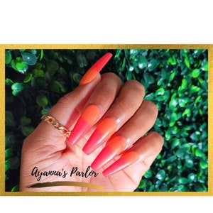 May include: Close-up of a hand with long, pink and orange ombre press-on nails. The nails are long and pointed, with a gradient of color from pink at the base to orange at the tip. The hand is wearing a gold chain ring.