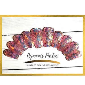 May include: A set of 10 pink press-on nails with iridescent glitter and a holographic finish. The nails are arranged in a semi-circle and have a rounded shape. The text "Ayanna's Parlor" and "Suggared Opals Press On Set" is visible below the nails.