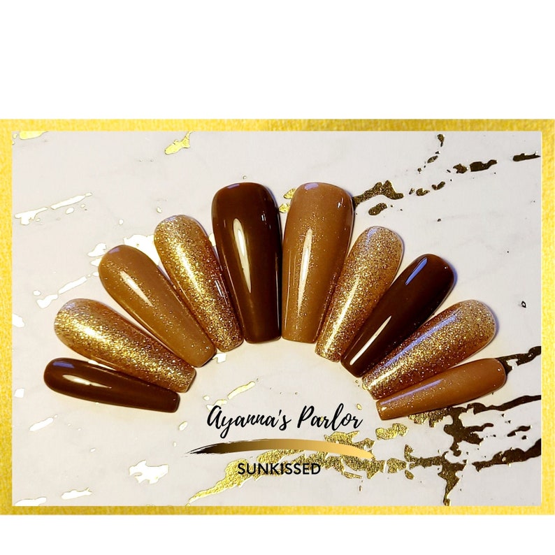 May include: A set of ten long, almond-shaped press-on nails in shades of brown and gold. Some nails have a glitter finish. The nails are arranged in a semi-circle on a white surface. The text "Ayanna's Parlor" and "Sunkissed" are visible below the nails.